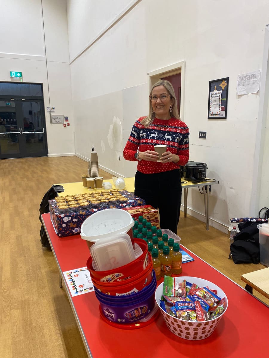 A volunteer in a Christmas jumper behind a table of mince pies, hot drinks and sweets